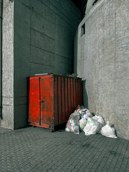 A large pile of black rubbish bags, most of which are tightly knotted and filled with waste, is stacked against a building with reflective tiled walls. Some of the bags appear to be bulging, indicatin