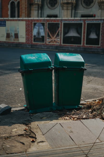 Two green plastic rubbish bins with closed lids positioned side by side on a pavement near a curb, with black clips securing the lids. The bins are made of durable plastic with a smooth, slightly refl