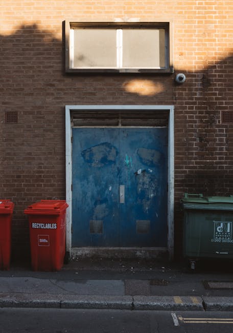 Two green plastic rubbish bins with closed lids positioned side by side on a pavement near a curb, with black clips securing the lids. The bins are made of durable plastic with a smooth, slightly refl