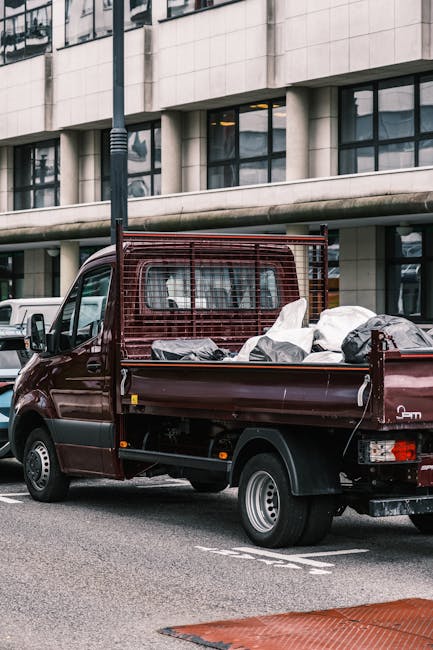 A small burgundy flatbed truck parked on a city street in front of a modern office building with large glass windows and concrete facade. The flatbed is loaded with various types of waste materials, i